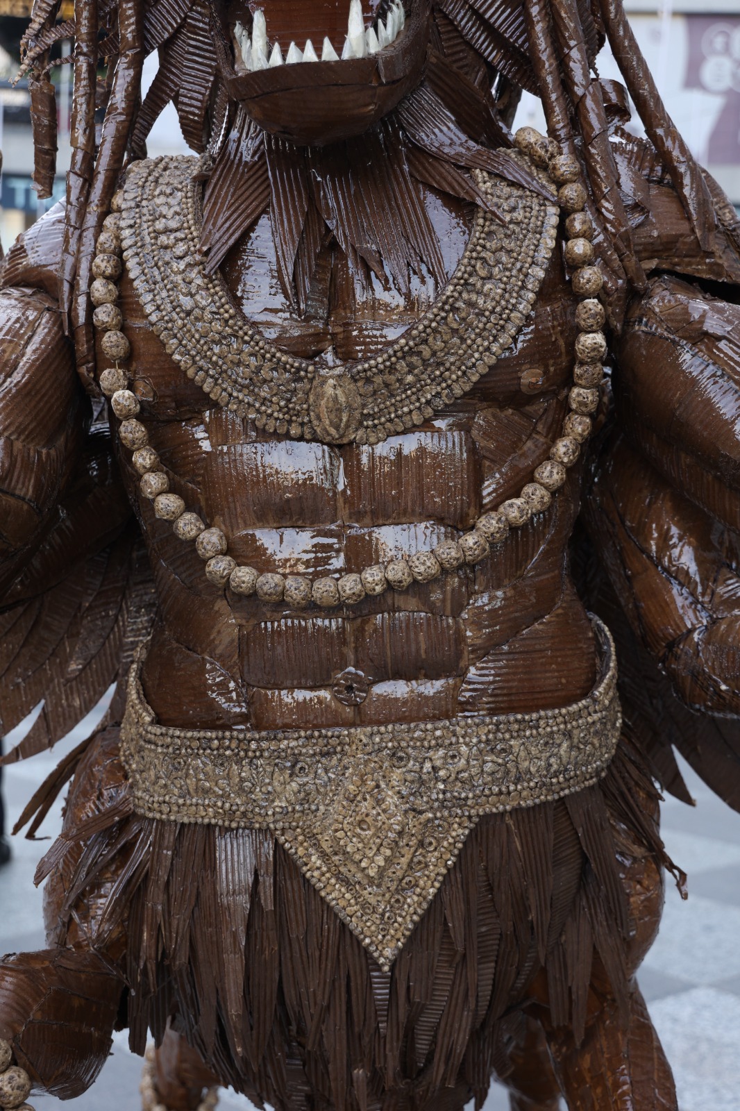 Detailed view of the layered corrugated cardboard torso, pectoral muscles, beaded garland necklace, and ornate carved belt.