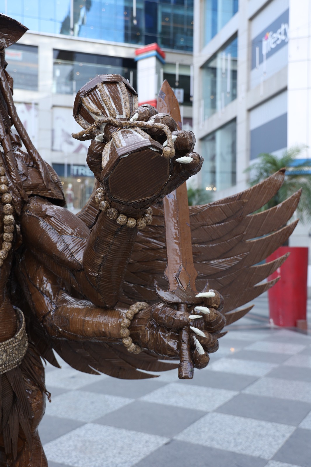 Close-up of the sculpture's left clawed hands holding a cardboard damru drum and sculpted sword with beaded bracelet.
